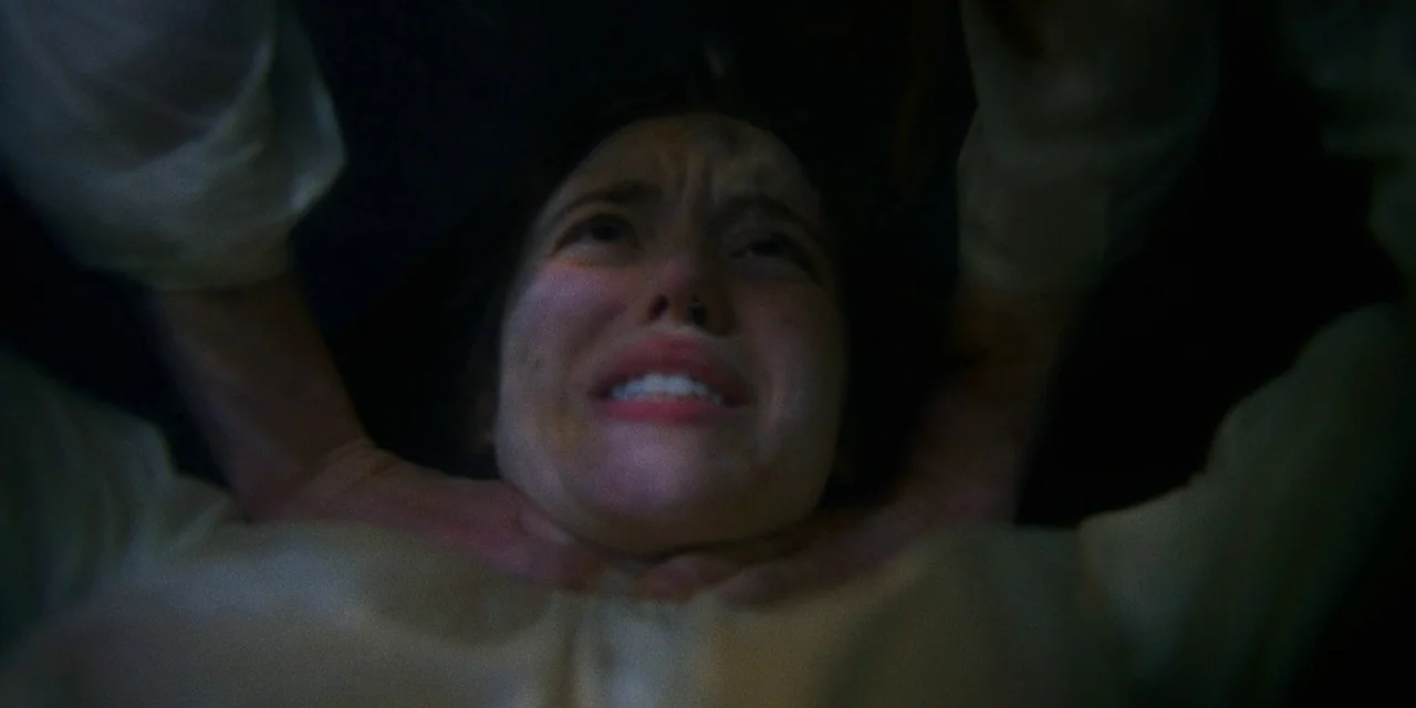 Immaculate (2024) movie still: closeup — A dimly lit, obscured environment surrounds a woman lying prone. Her face displays distress while hands…