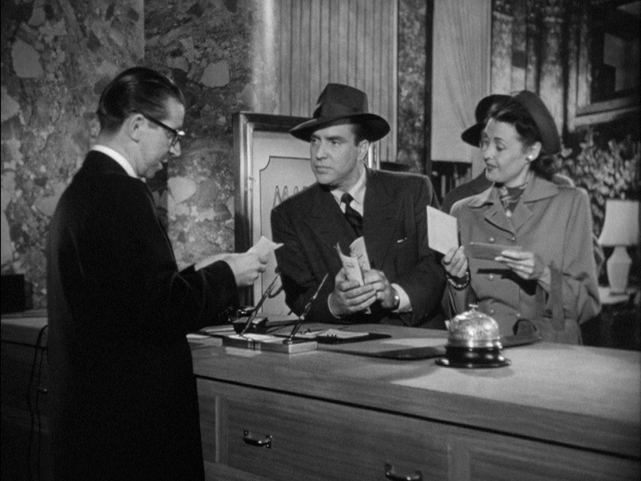D.O.A. (1949) movie still: medium — A polished hotel reception desk with a silver service bell sits in the foreground. A man and woman stand…
