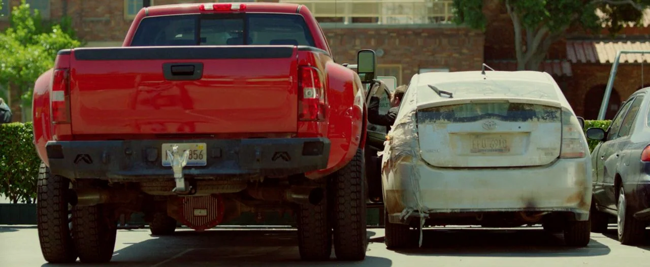 Cooties (2014) movie still: medium — A bright red dually pickup truck is parked next to a damaged white sedan. The sedan has a taped rear window and…
