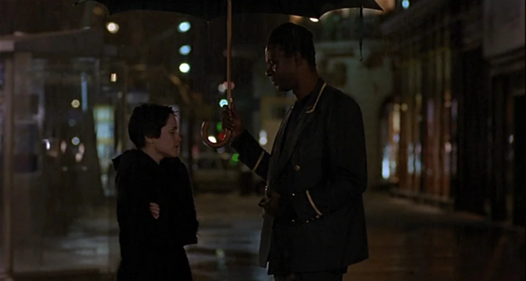 Trouble Every Day (2001) movie still: medium — A wet city sidewalk at night is illuminated by distant streetlights. A man holds an umbrella over a…