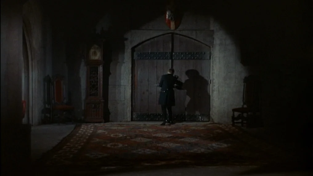 Targets (1968) movie still: wide — A dark hall features stone walls, a tall wooden door, and an ornate rug. A lone man in a dark coat reaches to…