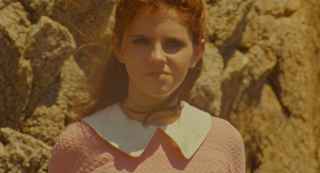 Moonrise Kingdom (2012) movie still: medium — A teenage girl stands outdoors with a textured rocky surface blurred in the background. She faces the…