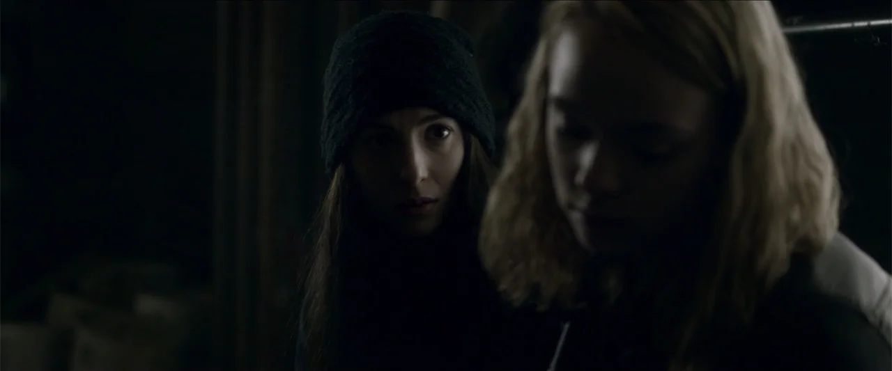 The Hollow Child (2018) movie still: medium — A dark indoor space with minimal light and obscured background details. Two young women occupy the…