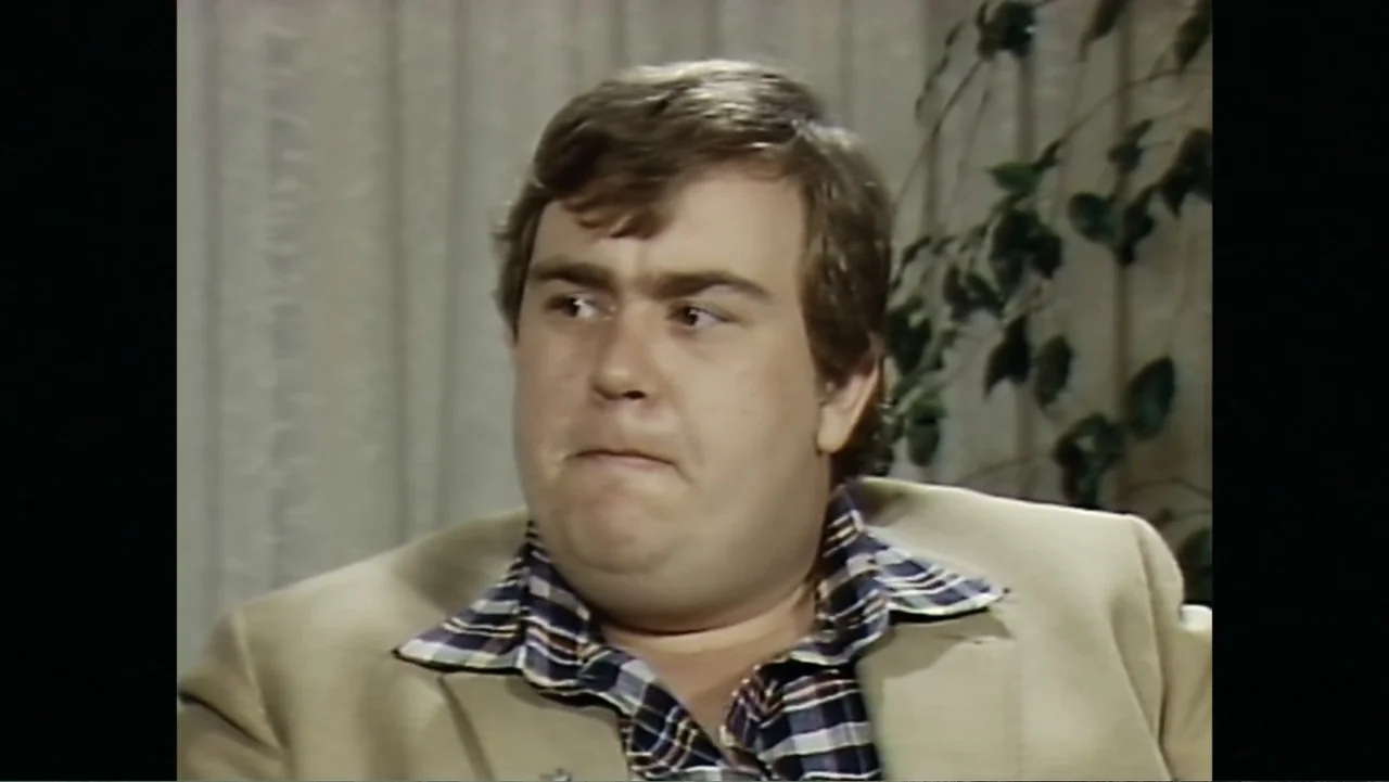 John Candy: I Like Me (2025) movie still: closeup — A man sits before a light-colored vertical blind backdrop with a plant visible behind. He looks…