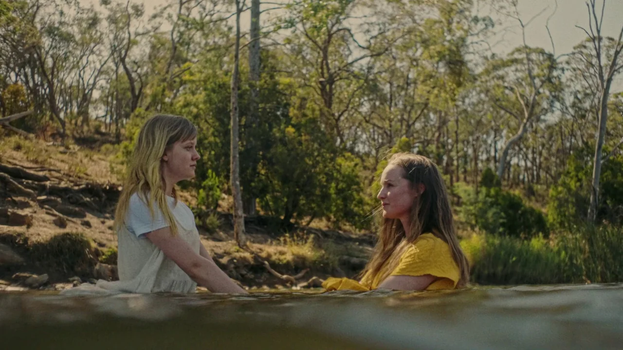 My First Summer (2020) movie still: medium — Two women stand waist-deep in a shallow creek surrounded by a wooded landscape. The setting features…