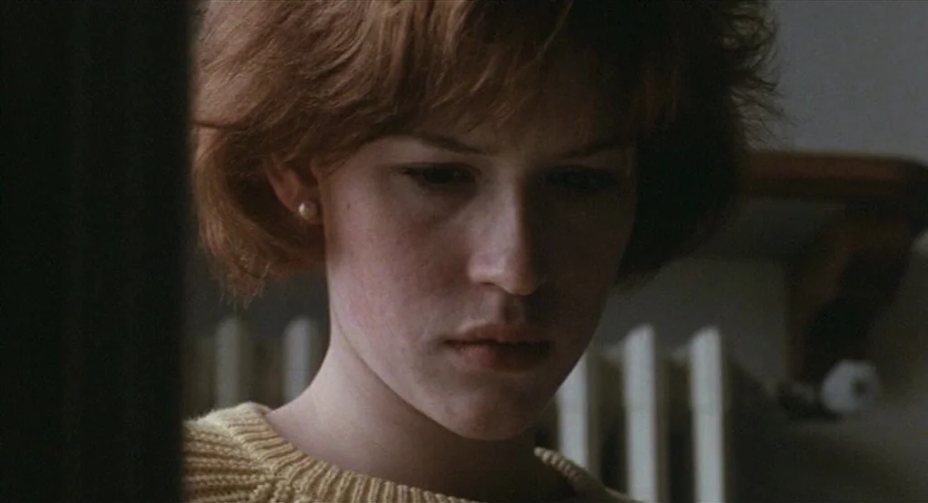 King Lear (1987) movie still: closeup — A close-up view of a young woman standing in a minimally furnished interior space. She gazes downward while…
