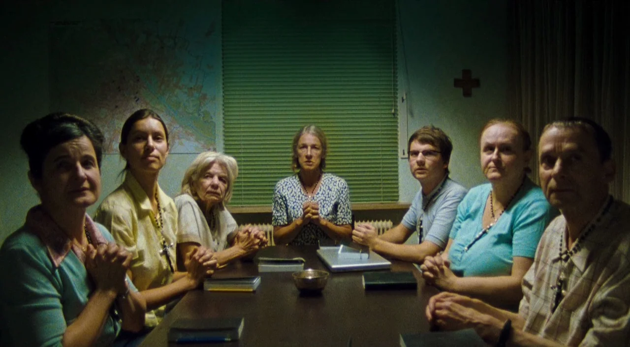 Paradise: Faith (2012) movie still: wide — A austere room with a map on the wall and green blinds over windows. Seven people sit in a line around a…