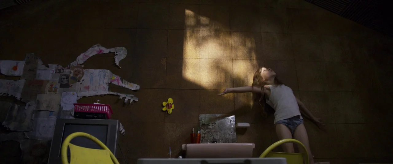 Room (2015) movie still: wide — A dark, cramped space has brown walls lined with children's drawings and a small sink. A young girl stands near the…