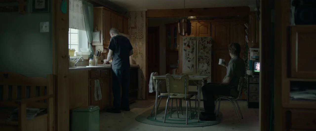 Small Crimes (2017) movie still: wide — A dated residential interior with wood cabinets and a linoleum floor. One man prepares food at the counter…