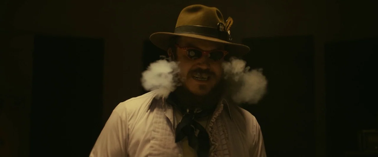 Blaze (2018) movie still: medium — A dimly lit interior space serves as the background for a man in a hat. He exhales thick clouds of smoke from his…