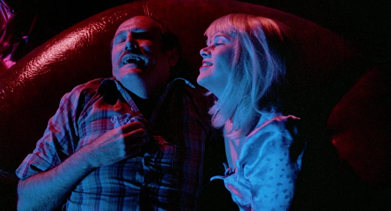Crimes of Passion (1984) movie still: medium — A dark booth with red vinyl upholstery serves as the backdrop for two people. A balding man with a…
