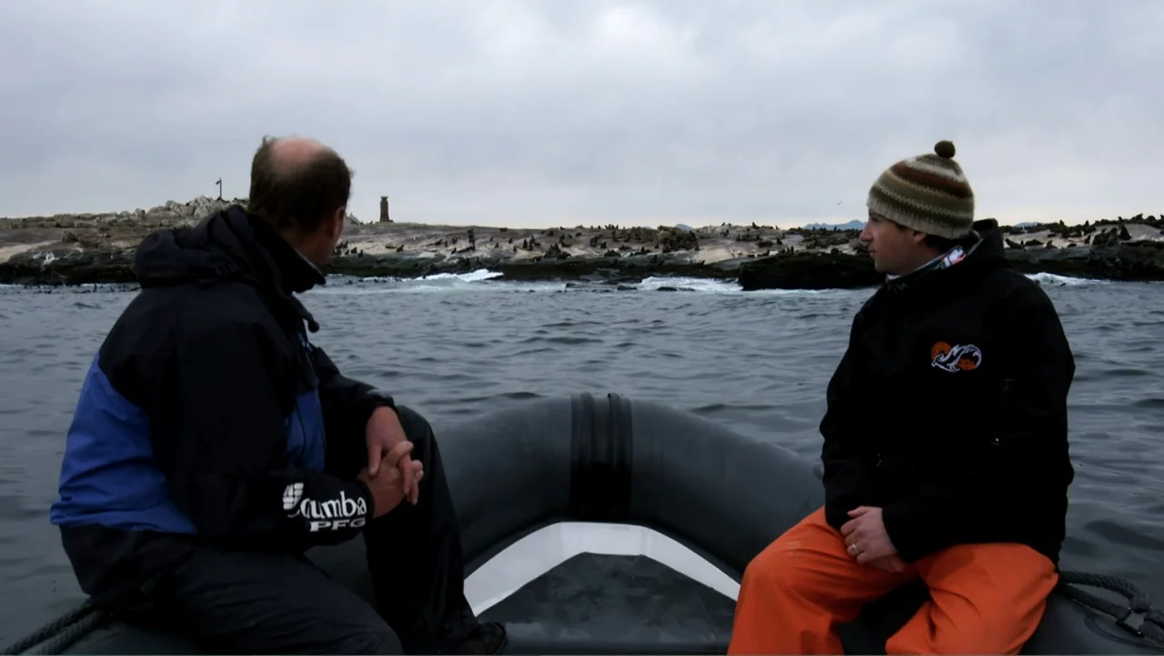 Air Jaws: Night Stalker (2016) movie still: medium — Two men in waterproof gear sit inside a black inflatable motorboat on open water. The shoreline…