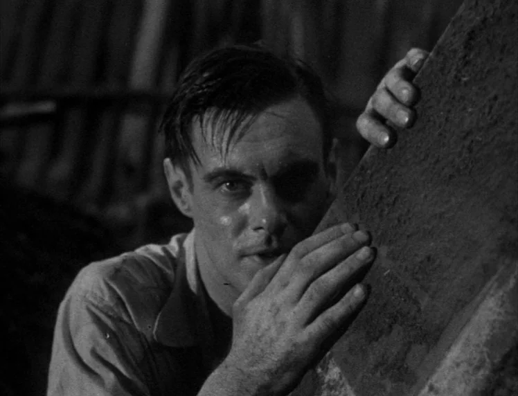 Frankenstein movie still: closeup — A man hides behind a rough stone block in a dark, outdoor environment. He leans forward with a hand pressed…