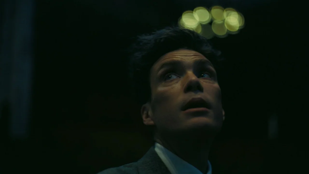 Oppenheimer (2023) movie still: closeup — A dark space features a man staring upward at an unseen light source. The background contains a soft,…