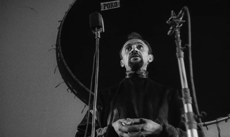 Man Is Not Bird (1965) movie still: medium — A dimly lit recording studio features a large black circular baffle behind the subject. A man in a dark…
