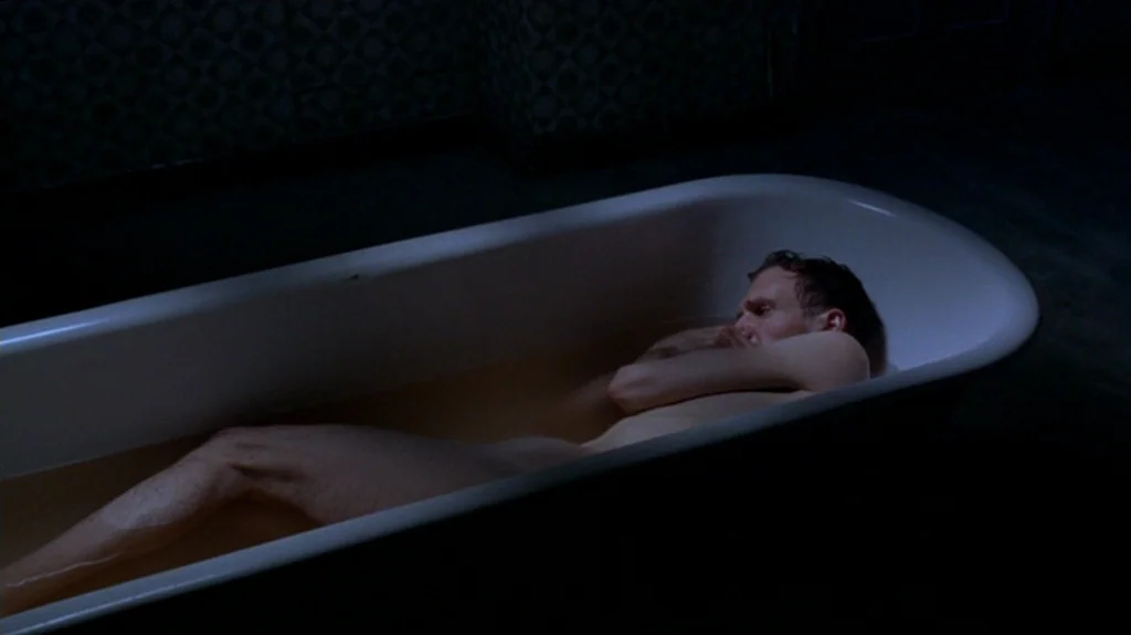 Spider (2002) movie still: medium — A dark, dimly lit room contains a white porcelain bathtub filled with water. A man lies curled up in the tub,…
