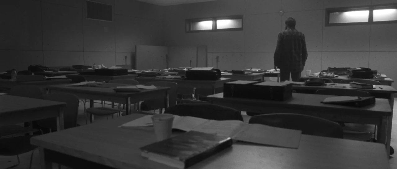 Polytechnique (2009) movie still: wide — A dark classroom with rows of wooden desks and scattered notebooks. A man stands alone by the back wall…