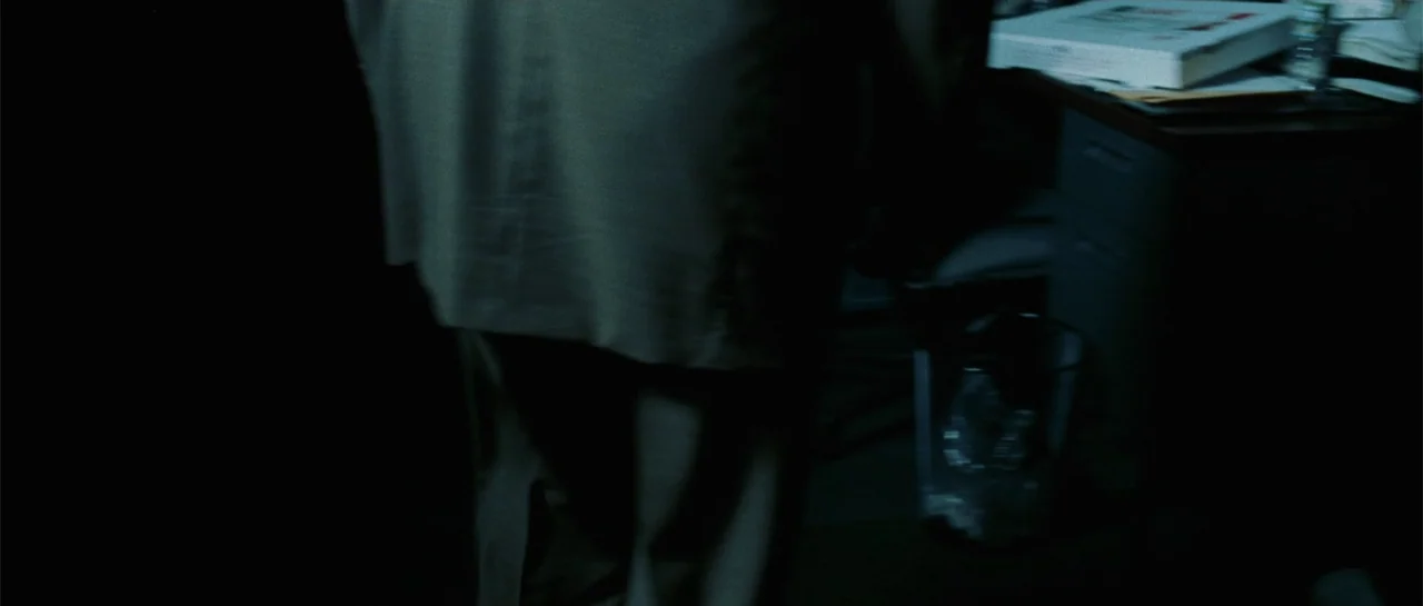 Inside Man (2006) movie still: medium — A dark office contains a desk with a pizza box and a wastebasket. A person in a light-colored jacket stands…