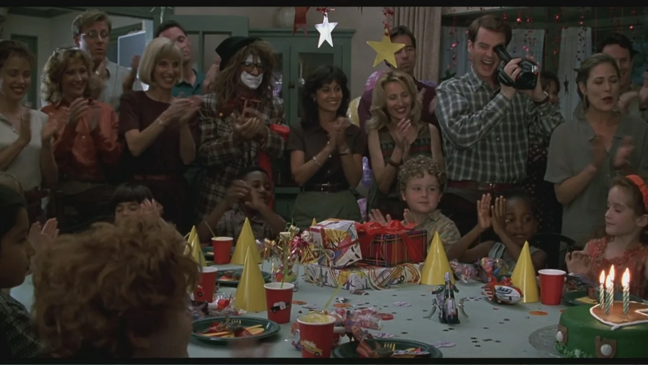 Liar Liar (1997) movie still: medium — A group of adults and children stand around a table decorated for a party. The group claps as a person behind…