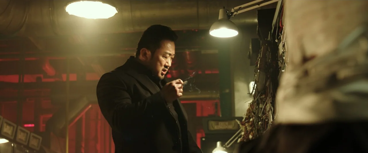 The Gangster, the Cop, the Devil (2019) movie still: medium — A dimly lit industrial workspace features exposed wiring and a desk lamp. A man stands…