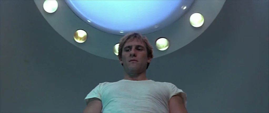 The Moon in the Gutter (1983) movie still: medium — A man stands beneath a large, circular ceiling light fixture in a sterile, modern room. He looks…