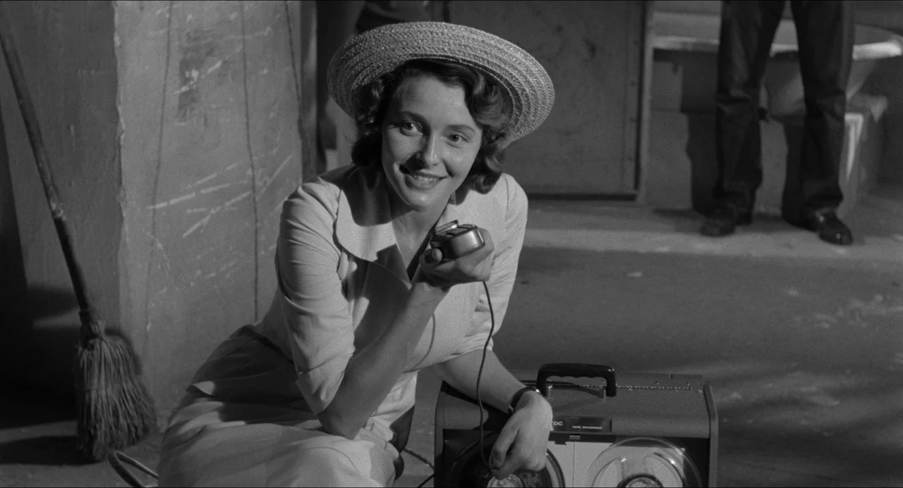 A Face In The Crowd (1957) movie still: medium — A woman in a light-colored dress and straw hat crouches in a dark alleyway. She holds a small…