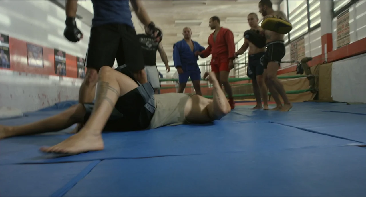 Boyka: Undisputed IV (2016) movie still: medium — A training mat in a gym features multiple men training in martial arts attire. A man rests on the…