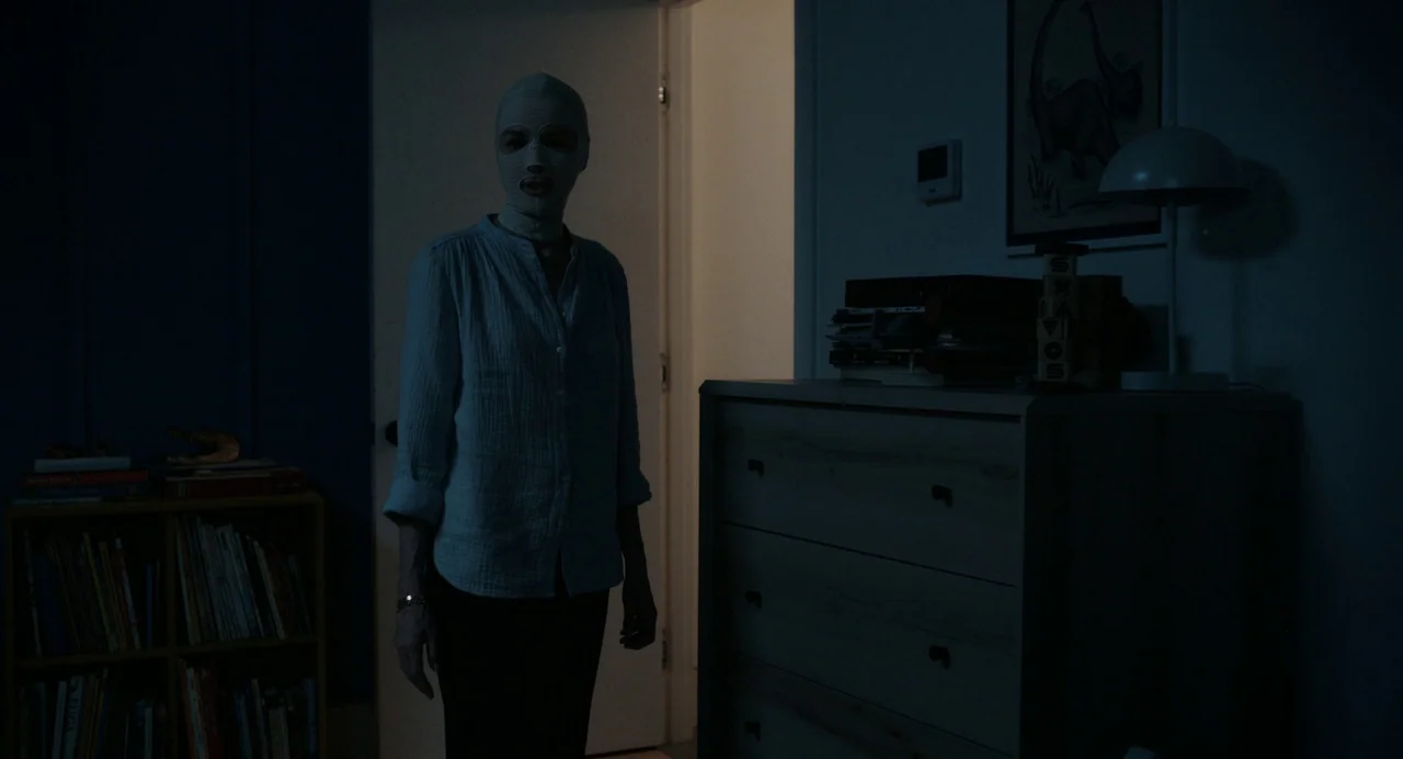Goodnight Mommy (2022) movie still: medium — A dimly lit room contains a wooden dresser and a low shelf full of books. A figure wearing a white…