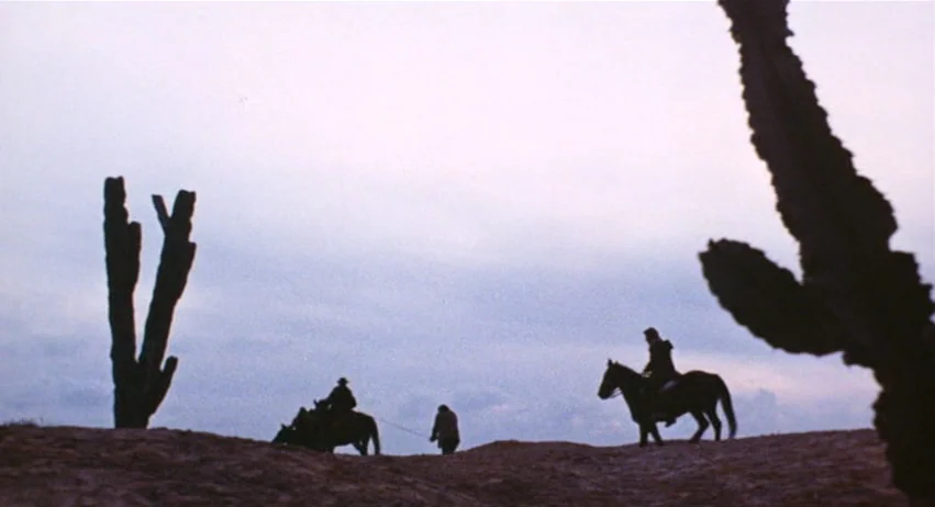 Roy Colt and Winchester Jack (1970) movie still: wide — A dry, rocky desert landscape silhouetted against a pale, fading evening sky. Three figures…