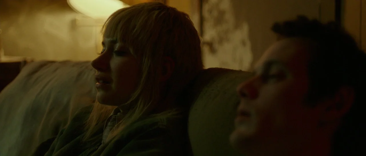 Green Room (2015) movie still: medium — A dark, dimly lit room contains two people sitting on a couch. The man and woman sit side by side with their…