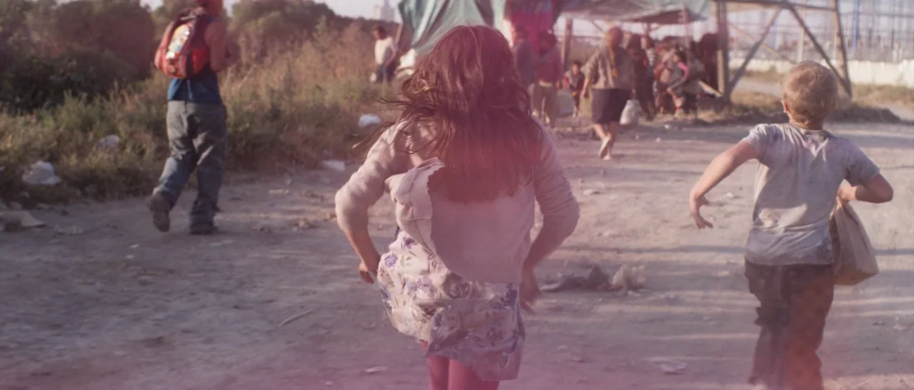 Elysium (2013) movie still: wide — A group of children runs along a dry, dusty track beside a makeshift camp. The ground is covered in light-colored…