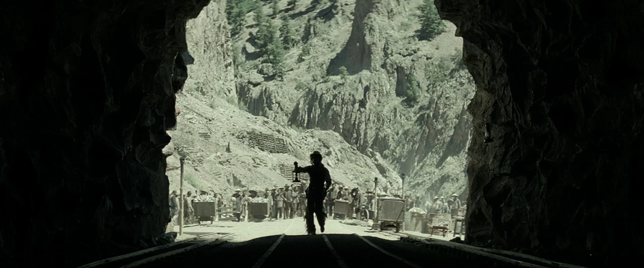 The Lone Ranger (2013) movie still: wide — A rugged mountain landscape features a dark rock tunnel framing a bustling mine site. A lone figure in…