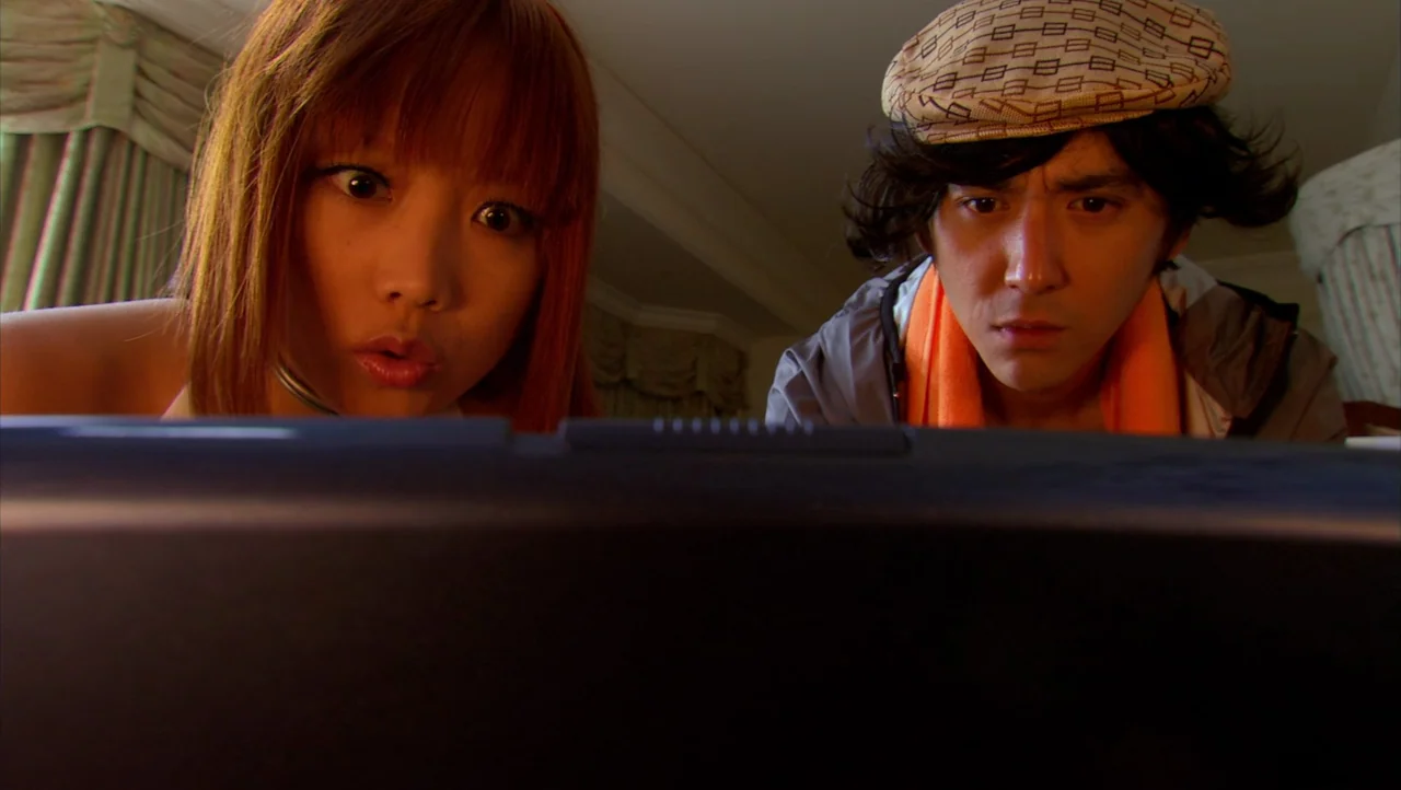 Cutie Honey (2004) movie still: medium — A dimly lit room with draped curtains visible in the blurred background. A young man and woman lean forward…