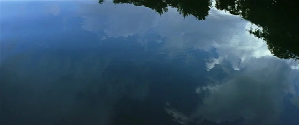 The New World (2005) movie still: establishing — The calm surface of a lake reflects white clouds and blue sky overhead. Dark silhouettes of trees…