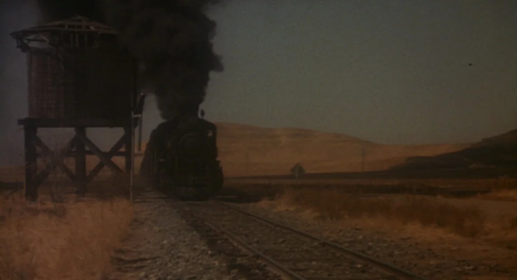 Bound For Glory (1976) movie still: wide — A dry, desolate landscape features railroad tracks passing a weathered wooden water tower. A dark steam…
