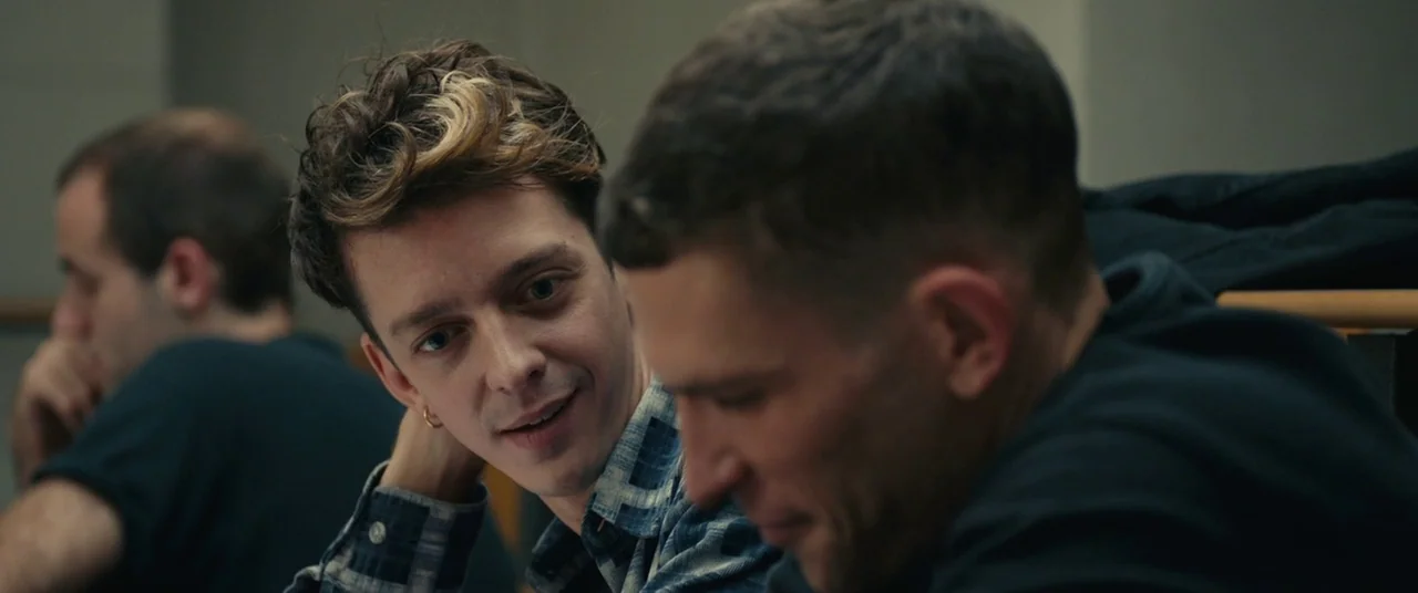 BPM (Beats Per Minute) (2017) movie still: medium — An indoor educational setting features rows of wooden desks and chairs. A young man in a plaid…