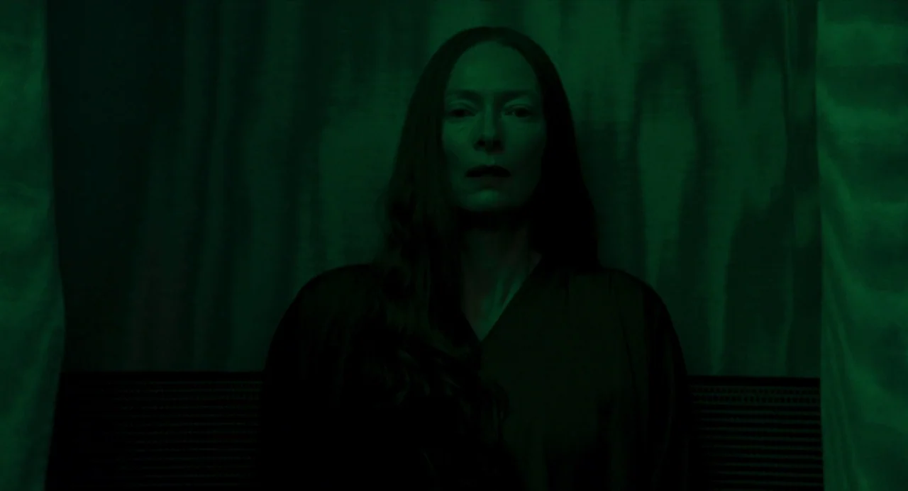Suspiria (2018) movie still: medium — A dark space features vertical fabric curtains acting as a backdrop behind the subject. A woman sits centrally…
