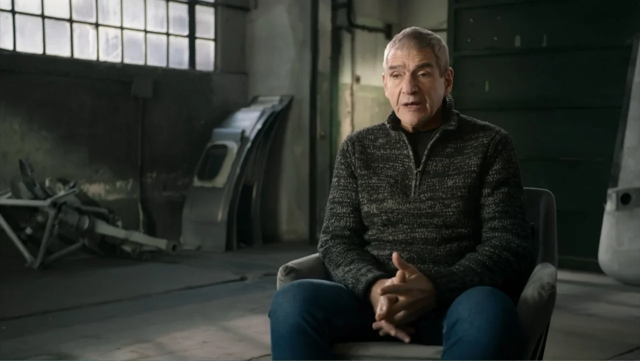Transfers (2024) movie still: medium — An industrial warehouse space with concrete floors and large paned windows in the background. A man sits in a…