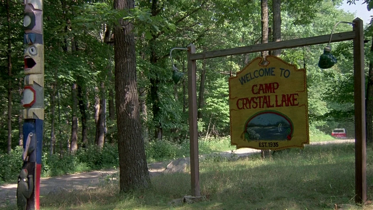 Friday the 13th (1980) movie still: establishing — A wooden sign hangs from a metal frame near a dirt road in woods. The sign displays the text…