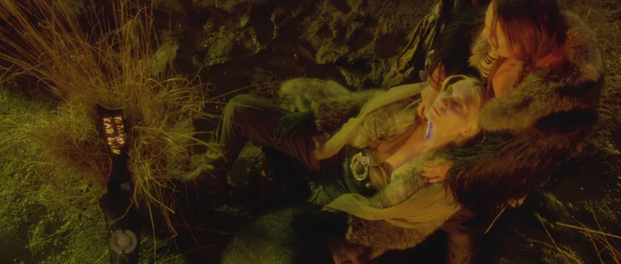 After Blue (2021) movie still: medium — A dark, muddy marsh at night with tall, dead stalks of grass nearby. One person sits in the mud, cradling a…