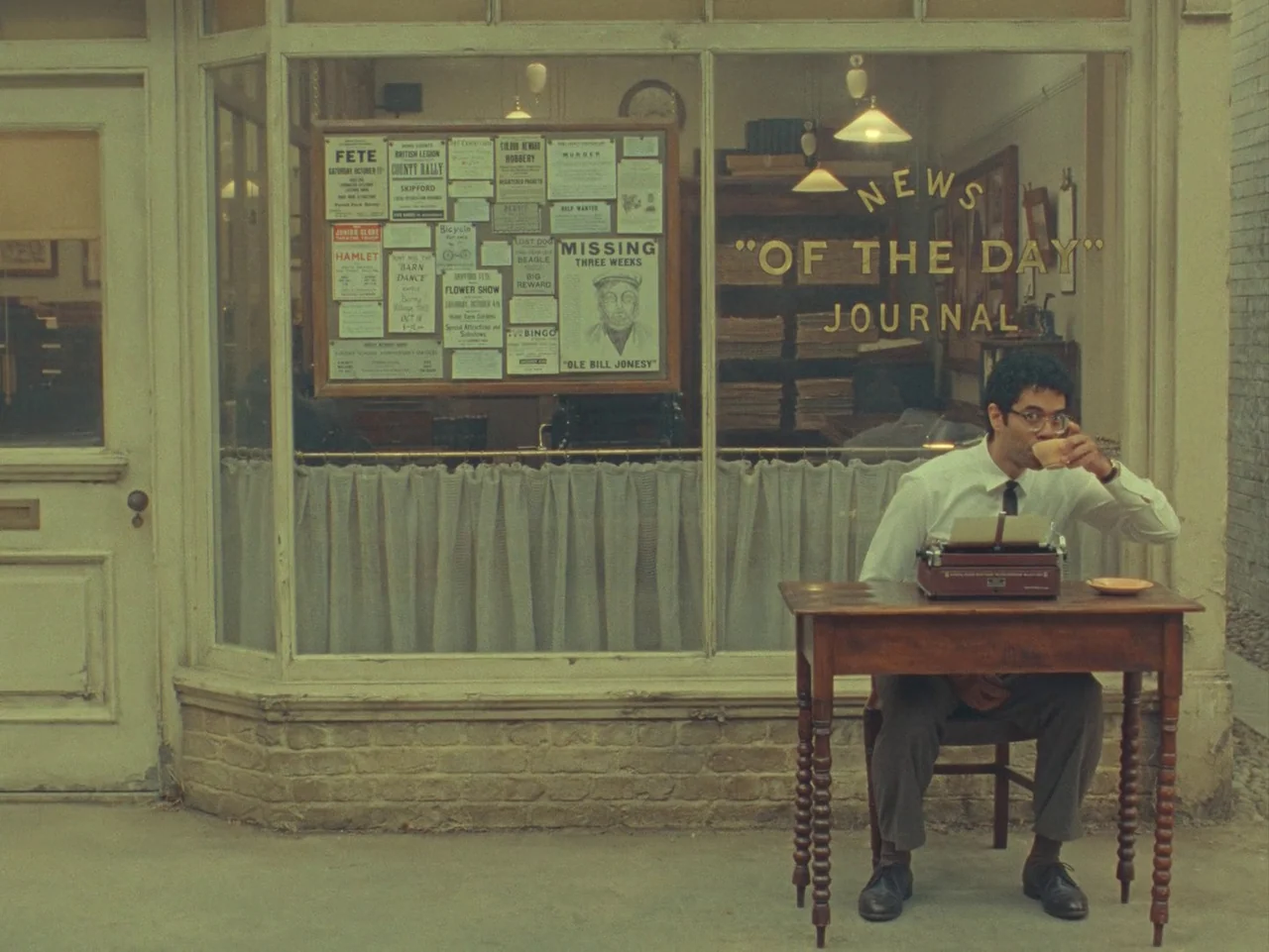 The Rat Catcher (2023) movie still: wide — The exterior of a vintage newspaper shop features a large window with posters displayed. A man sits at a…