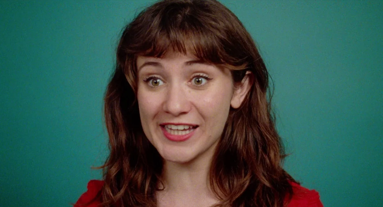 Mr. Roosevelt (2017) movie still: closeup — A woman stands centered against a solid teal-colored studio backdrop. She smiles directly toward the…