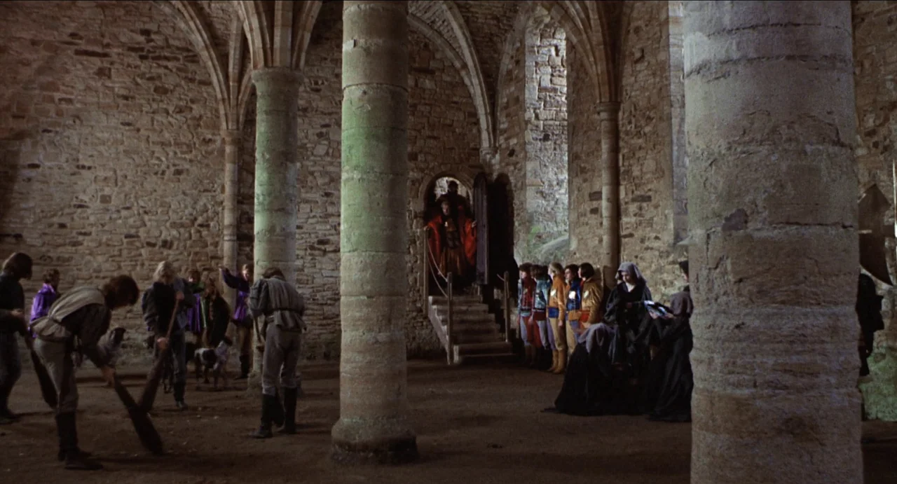 The Canterbury Tales (1972) movie still: wide — A stone vaulted interior with thick columns and rough masonry walls shows activity. Figures dressed…
