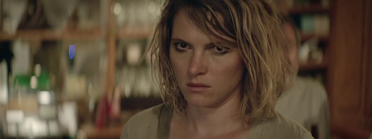 Entertainment (2015) movie still: closeup — A dimly lit room with blurred shelves and household objects in the background. A woman with messy hair…