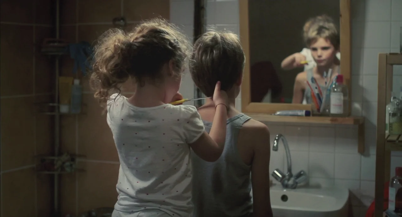 Tomboy (2011) movie still: medium — A modest home bathroom features white tiled walls and a wooden framed vanity mirror. A young girl uses scissors…