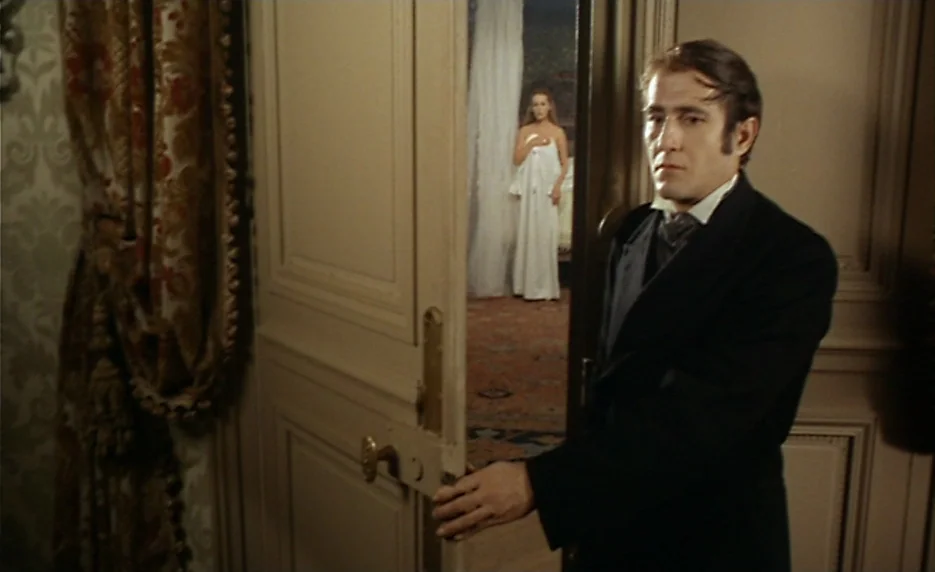 The Immortal Story (1968) movie still: medium — A man stands in an ornate hallway before an open wood-paneled door. Through the doorway, a woman in…
