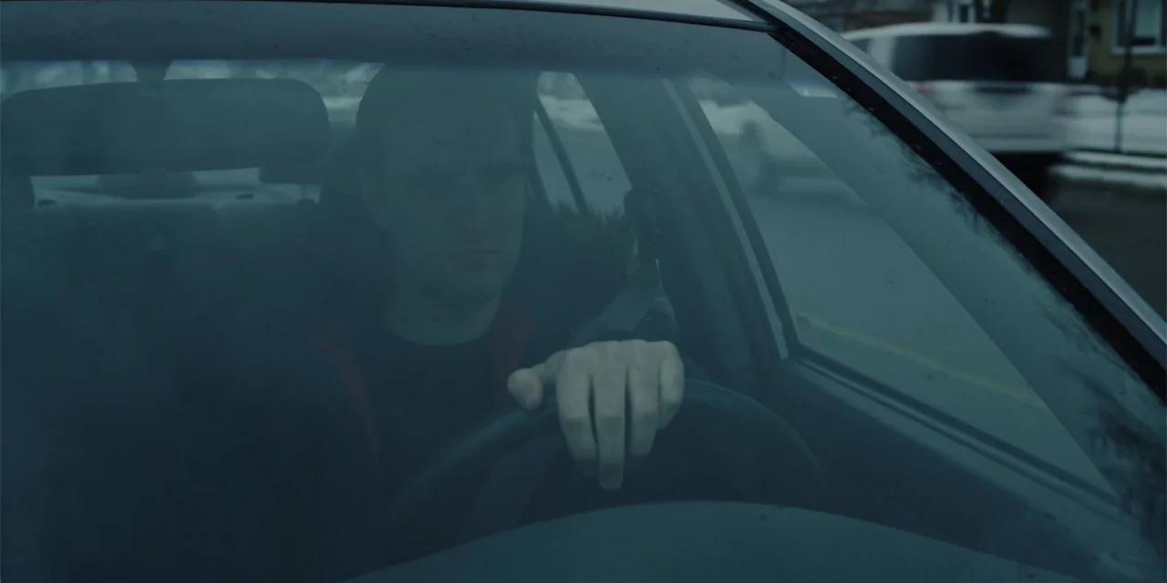 Lobos (2019) movie still: medium — The interior of a parked car shows a view through the front windshield. A man sits in the driver seat holding the…