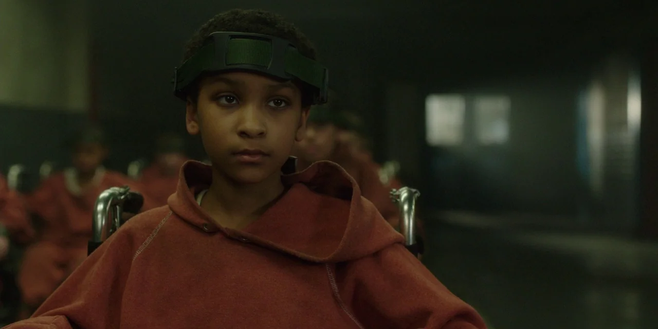 The Girl With All The Gifts (2016) movie still: closeup — A dark, dimly lit institutional hall contains several children sitting in chairs in rows.…