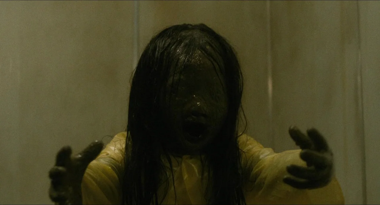 Dark Water (2002) movie still: closeup — A figure stands inside a dark, tiled enclosure with beige walls. The figure wears a wet yellow poncho and…