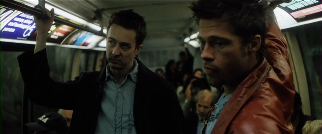 Fight Club (1999) movie still: medium — The interior of a dimly lit subway car shows advertisements along the ceiling panels. Two men stand together…